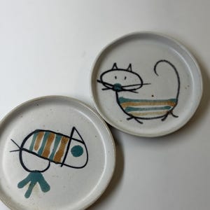 May include: Two round, off-white ceramic plates featuring hand-drawn illustrations in black, teal, and mustard yellow. One plate displays a stylized cat with striped markings, while the other shows a geometric creature with a circular eye and tail.