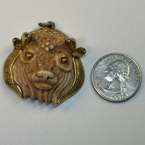 May include: A vintage pendant featuring a carved bison head with a gold-tone frame. The pendant has a textured, off-white finish with gold-colored accents. A U.S. quarter is next to the pendant for size comparison.