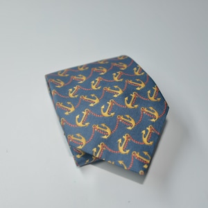 Pode incluir: Uma gravata azul dobrada com um padr&atilde;o repetido de &acirc;ncoras douradas conectadas por correntes vermelhas. A gravata &eacute; feita de um tecido liso e &eacute; adequada para ocasi&otilde;es formais ou semi-formais.