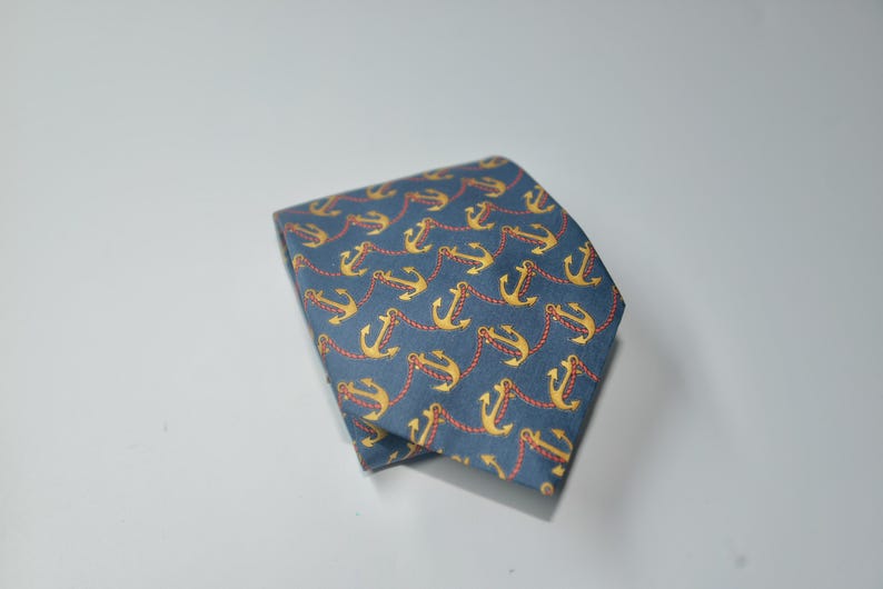 Pode incluir: Uma gravata azul dobrada com um padr&atilde;o repetido de &acirc;ncoras douradas conectadas por correntes vermelhas. A gravata &eacute; feita de um tecido liso e &eacute; adequada para ocasi&otilde;es formais ou semi-formais.