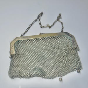 May include: A vintage silver mesh purse with a chain handle. The purse has a rectangular frame and a delicate, woven mesh body. The chain handle is also silver, and the purse has a small, decorative bead at the bottom.