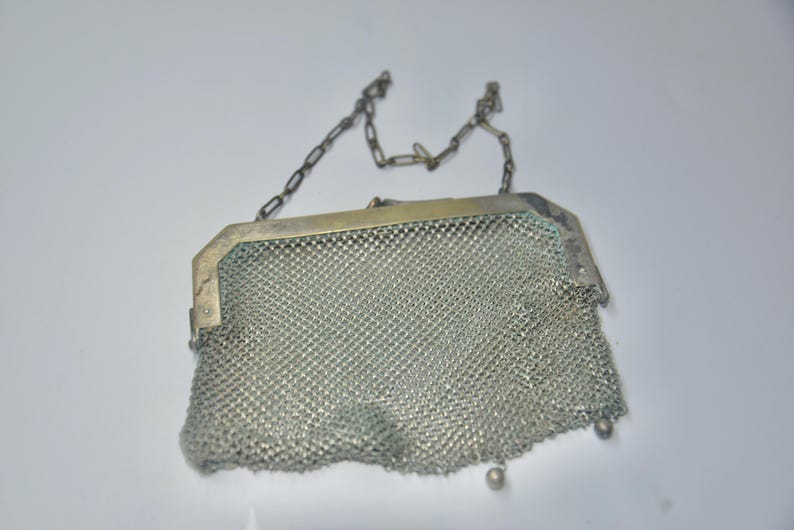 May include: A vintage silver mesh purse with a chain handle. The purse has a rectangular frame and a delicate, woven mesh body. The chain handle is also silver, and the purse has a small, decorative bead at the bottom.