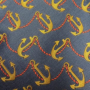 Pode incluir: Close-up de um tecido azul marinho com um padr&atilde;o repetido de &acirc;ncoras douradas ligadas por corda vermelha. As &acirc;ncoras t&ecirc;m um design cl&aacute;ssico, e a corda adiciona um tema n&aacute;utico. O tecido tem uma textura subtil.