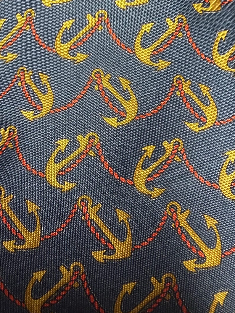 Pode incluir: Close-up de um tecido azul marinho com um padr&atilde;o repetido de &acirc;ncoras douradas ligadas por corda vermelha. As &acirc;ncoras t&ecirc;m um design cl&aacute;ssico, e a corda adiciona um tema n&aacute;utico. O tecido tem uma textura subtil.