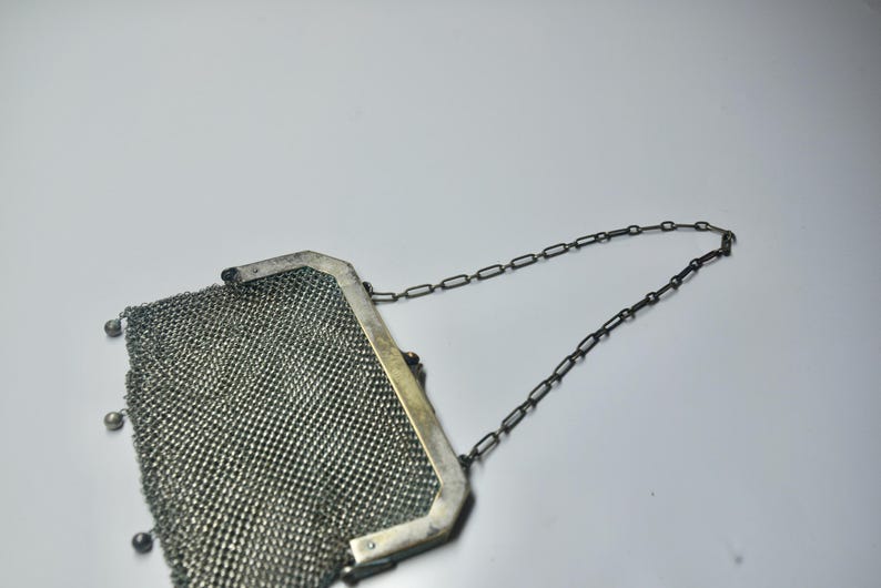 May include: A vintage silver mesh purse with a chain handle. The purse has a rectangular frame and small decorative beads at the bottom. The mesh is tightly woven, and the chain is dark in color.