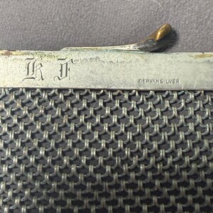 May include: Close-up of a vintage silver mesh purse. The purse features a metal frame with the text "GERMAN SILVER" and the initials "I K" engraved. The clasp has a gold-toned finish. The mesh is a tightly woven pattern.
