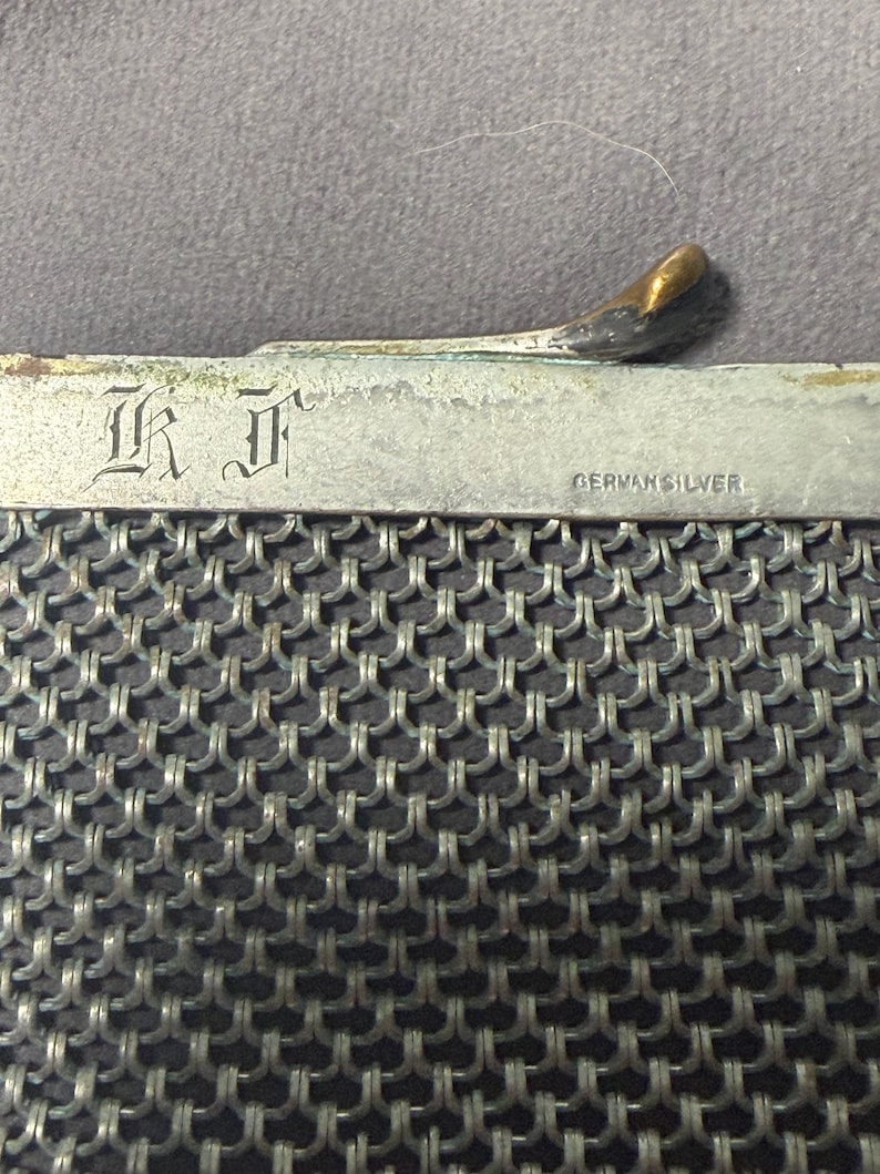 May include: Close-up of a vintage silver mesh purse. The purse features a metal frame with the text "GERMAN SILVER" and the initials "I K" engraved. The clasp has a gold-toned finish. The mesh is a tightly woven pattern.