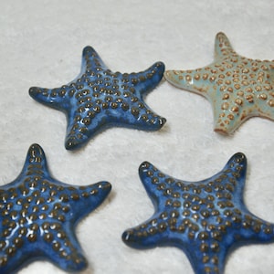 May include: Four ceramic starfish decorations. Three are a deep blue with brown accents, and one is a light blue with brown accents. The starfish have a textured surface and are arranged on a white background. These decorative items are suitable for coastal-themed decor.