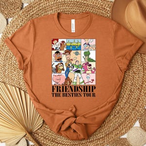 Puede incluir: Una camiseta naranja con un gr&aacute;fico colorido de personajes de la pel&iacute;cula animada Toy Story. El gr&aacute;fico est&aacute; dividido en cuadrados e incluye el texto "FRIENDSHIP THE BESTIES TOUR". La camiseta est&aacute; doblada y colocada sobre una superficie tejida.