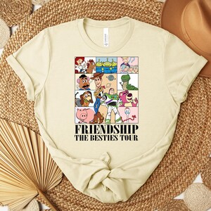 Puede incluir: Camiseta de color crema con un gr&aacute;fico colorido de personajes de la pel&iacute;cula Toy Story. El gr&aacute;fico est&aacute; dividido en cuadrados e incluye el texto "FRIENDSHIP THE BESTIES TOUR" en negro. La camiseta tiene cuello redondo y mangas cortas.