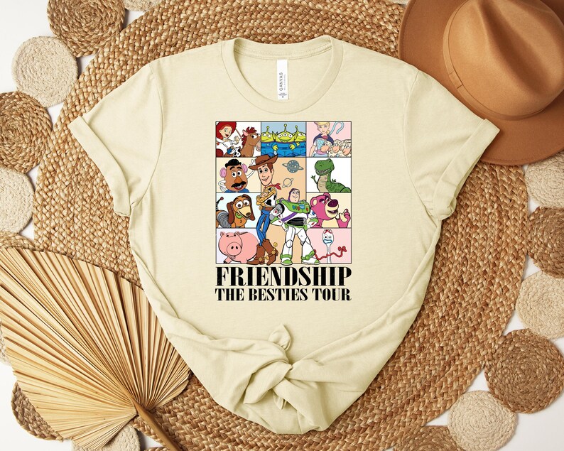 Puede incluir: Camiseta de color crema con un gr&aacute;fico colorido de personajes de la pel&iacute;cula Toy Story. El gr&aacute;fico est&aacute; dividido en cuadrados e incluye el texto "FRIENDSHIP THE BESTIES TOUR" en negro. La camiseta tiene cuello redondo y mangas cortas.