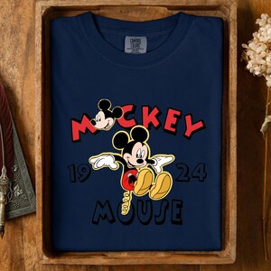 May include: Navy blue t-shirt with a Mickey Mouse graphic. The design includes the word "Mickey" in red, the year "1924", and the word "Mouse". The shirt is displayed in a wooden tray.