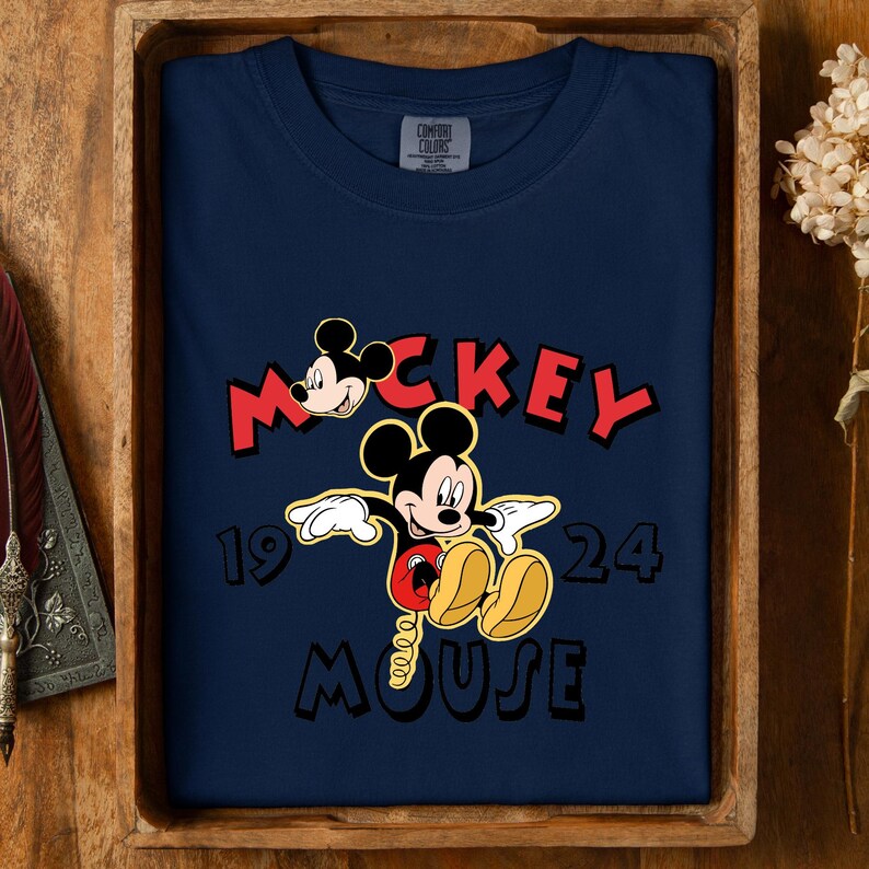 May include: Navy blue t-shirt with a Mickey Mouse graphic. The design includes the word "Mickey" in red, the year "1924", and the word "Mouse". The shirt is displayed in a wooden tray.