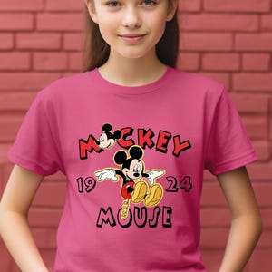 May include: A magenta t-shirt with a Mickey Mouse graphic. The design features the words "Mickey Mouse" in black and red, and the numbers "19" and "24". The t-shirt is being worn by a person.
