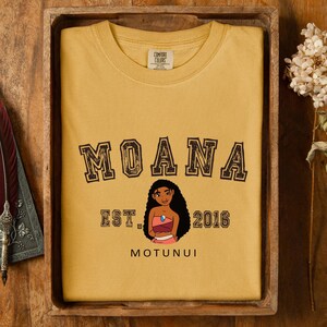 Pu&ograve; includere: Felpa girocollo giallo senape con la scritta "MOANA" in un font invecchiato. Sotto la parola "MOANA" c'&egrave; un'illustrazione di un personaggio dei cartoni animati e la scritta "EST. 2016 MOTUNUI". La felpa &egrave; piegata ordinatamente in un vassoio di legno.