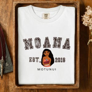 Pu&ograve; includere: T-shirt bianca con la scritta "MOANA" in marrone e bordata. Sotto, un'illustrazione di un personaggio dei cartoni animati, la scritta "EST. 2016" e la parola "MOTUNUI". La maglietta &egrave; esposta su un vassoio di legno.