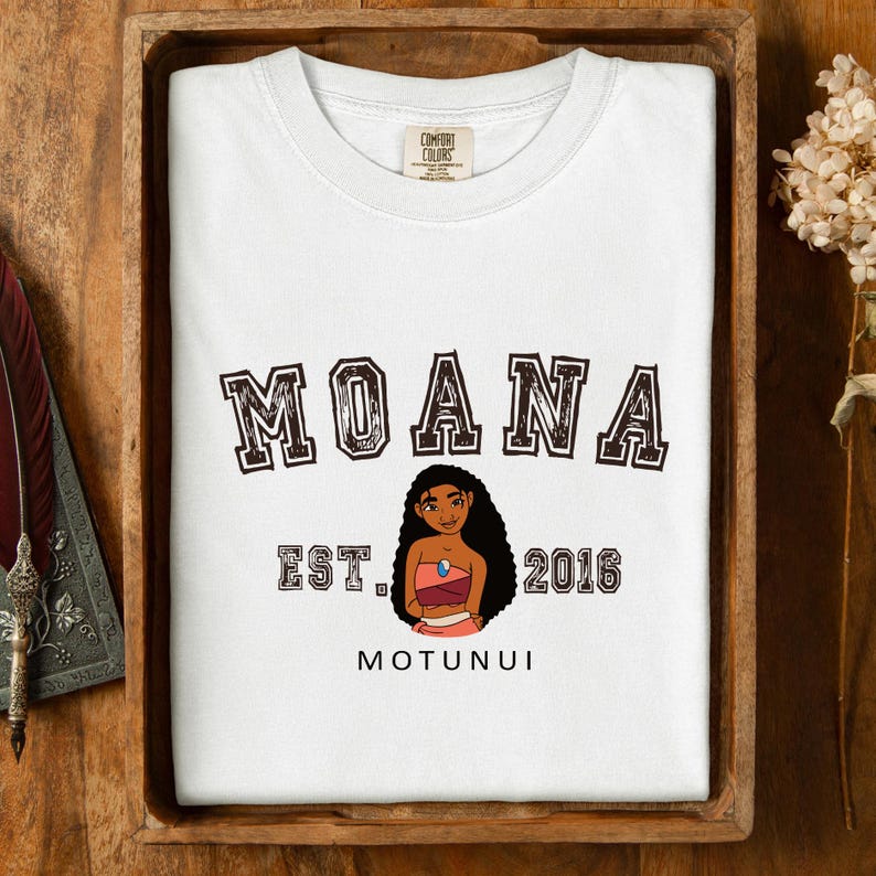 Pu&ograve; includere: T-shirt bianca con la scritta "MOANA" in marrone e bordata. Sotto, un'illustrazione di un personaggio dei cartoni animati, la scritta "EST. 2016" e la parola "MOTUNUI". La maglietta &egrave; esposta su un vassoio di legno.