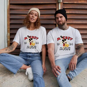 May include: Two people wearing white t-shirts featuring a Mickey Mouse graphic. The design includes the text "Mickey Mouse 1924" in red and black. They are both wearing blue jeans and are seated.