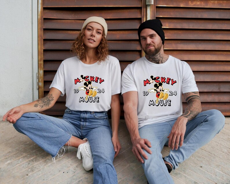 May include: Two people wearing white t-shirts featuring a Mickey Mouse graphic. The design includes the text "Mickey Mouse 1924" in red and black. They are both wearing blue jeans and are seated.