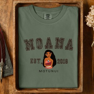 Pu&ograve; includere: T-shirt verde oliva con la scritta "MOANA" in lettere marroni. Sotto, un'illustrazione a cartoni animati di una donna in abito rosa e rosso. Il testo "EST. 2016" e "MOTUNUI" sono stampati sulla maglietta.