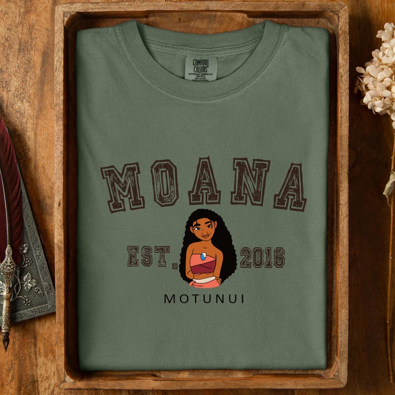 Pu&ograve; includere: T-shirt verde oliva con la scritta "MOANA" in lettere marroni. Sotto, un'illustrazione a cartoni animati di una donna in abito rosa e rosso. Il testo "EST. 2016" e "MOTUNUI" sono stampati sulla maglietta.