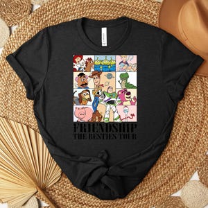 Puede incluir: Camiseta negra con una cuadr&iacute;cula de personajes de dibujos animados de Toy Story. El texto "FRIENDSHIP THE BESTIES TOUR" est&aacute; impreso debajo. La camiseta se muestra sobre una superficie tejida, con un sombrero y elementos decorativos.