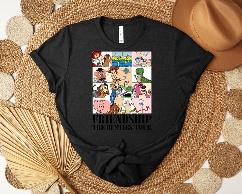 Puede incluir: Camiseta negra con una cuadr&iacute;cula de personajes de dibujos animados de Toy Story. El texto "FRIENDSHIP THE BESTIES TOUR" est&aacute; impreso debajo. La camiseta se muestra sobre una superficie tejida, con un sombrero y elementos decorativos.