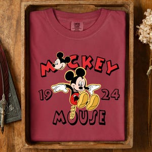 May include: A maroon Comfort Colors t-shirt with a Mickey Mouse graphic and the text "Mickey Mouse 1924". The design shows Mickey's face and full body in a classic cartoon style. The shirt is folded neatly in a wooden tray.