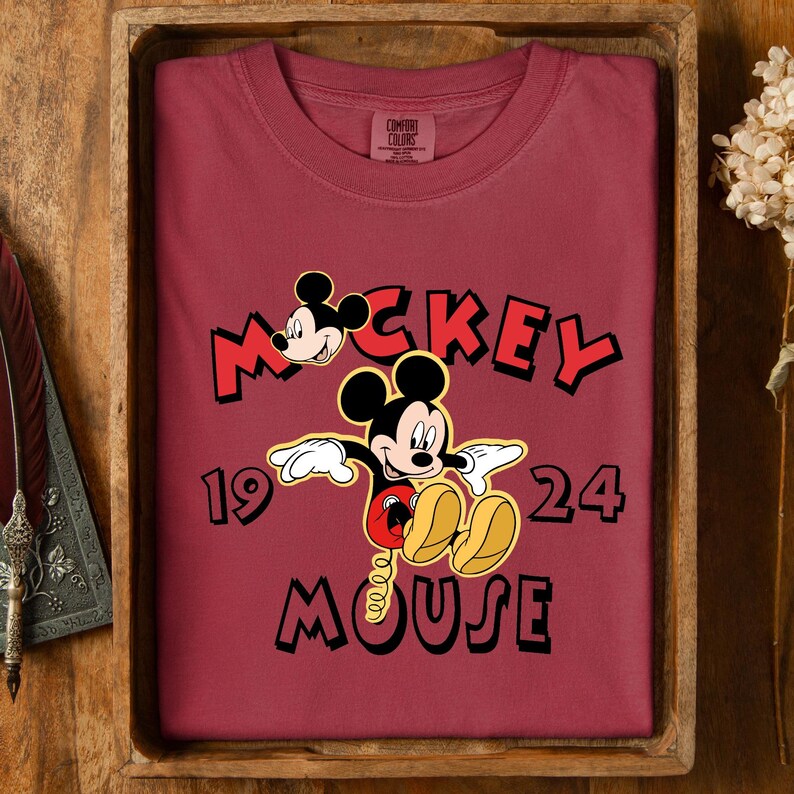 May include: A maroon Comfort Colors t-shirt with a Mickey Mouse graphic and the text "Mickey Mouse 1924". The design shows Mickey's face and full body in a classic cartoon style. The shirt is folded neatly in a wooden tray.