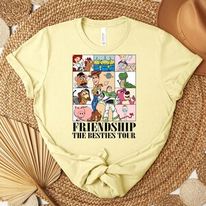 Puede incluir: Una camiseta amarilla clara con un gr&aacute;fico colorido de personajes de Toy Story. El gr&aacute;fico est&aacute; dividido en nueve cuadrados e incluye el texto "FRIENDSHIP THE BESTIES TOUR".