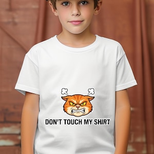 May include: White t-shirt featuring a cartoon cat with an angry expression in orange and yellow. The text "DON'T TOUCH MY SHIRT" is printed below the graphic in black, bold letters. The shirt is worn by a child.