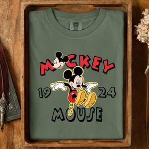 May include: An olive green t-shirt with a Mickey Mouse graphic and the text "Mickey Mouse 1924". The design shows Mickey's face and full body in a classic pose. The shirt is folded and presented in a wooden tray.