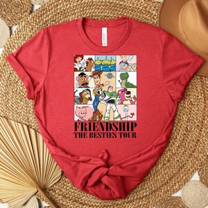 Puede incluir: Camiseta roja con un dise&ntilde;o gr&aacute;fico colorido de personajes de dibujos animados. El dise&ntilde;o est&aacute; dividido en cuadrados y contiene el texto "FRIENDSHIP THE BESTIES TOUR". La camiseta est&aacute; hecha de un material suave y tiene un cuello redondo cl&aacute;sico.
