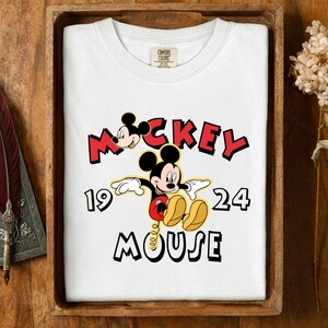 May include: White t-shirt with a Mickey Mouse graphic and the text "Mickey Mouse 1924" in red and black. The shirt is presented in a wooden tray.