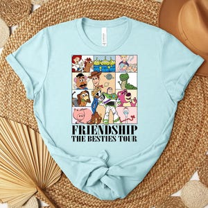 Puede incluir: Camiseta azul claro con un dise&ntilde;o de personajes de dibujos animados en una cuadr&iacute;cula. El texto "FRIENDSHIP THE BESTIES TOUR" est&aacute; impreso debajo. La camiseta es de material suave y tiene cuello redondo. Una prenda casual.