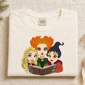 May include: A cream-colored t-shirt with a Comfort Colors label at the neck, featuring a graphic print of three cartoon characters with distinct hair colors—blonde, orange, and black—reading an open book together.