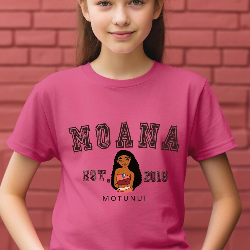Pu&ograve; includere: Una maglietta rosa con la scritta "MOANA" in nero, con contorno. Sotto, un'illustrazione di un personaggio dei cartoni animati, con il testo "EST. 2016" e "MOTUNUI". La maglietta &egrave; indossata da una persona.