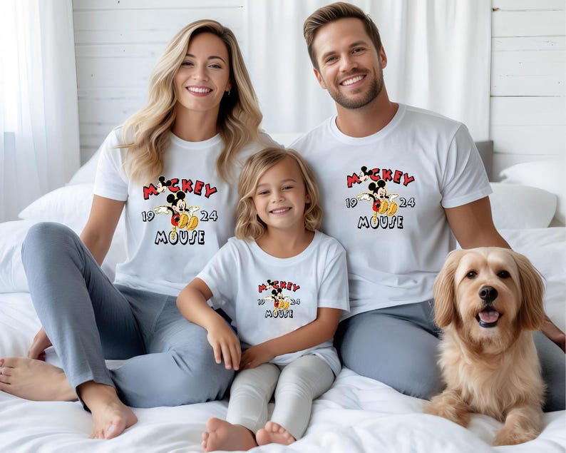 May include: A family and a dog are sitting on a bed, all wearing matching white t-shirts. The shirts feature a Mickey Mouse graphic with the words "Mickey Mouse" and the numbers "1924". The dog is a golden retriever.