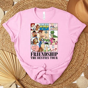 Puede incluir: Camiseta rosa claro con un dise&ntilde;o gr&aacute;fico colorido de personajes de dibujos animados en una cuadr&iacute;cula. El texto "FRIENDSHIP THE BESTIES TOUR" est&aacute; impreso debajo. La camiseta est&aacute; hecha de un material suave.