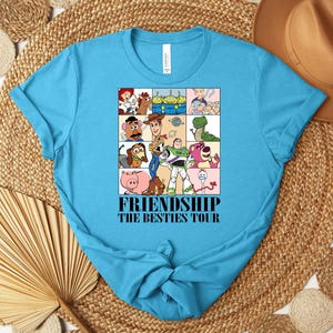 Puede incluir: Una camiseta turquesa con un gr&aacute;fico colorido de personajes de dibujos animados en una cuadr&iacute;cula. El gr&aacute;fico incluye personajes como Woody y Buzz Lightyear. El texto "FRIENDSHIP THE BESTIES TOUR" est&aacute; impreso debajo.