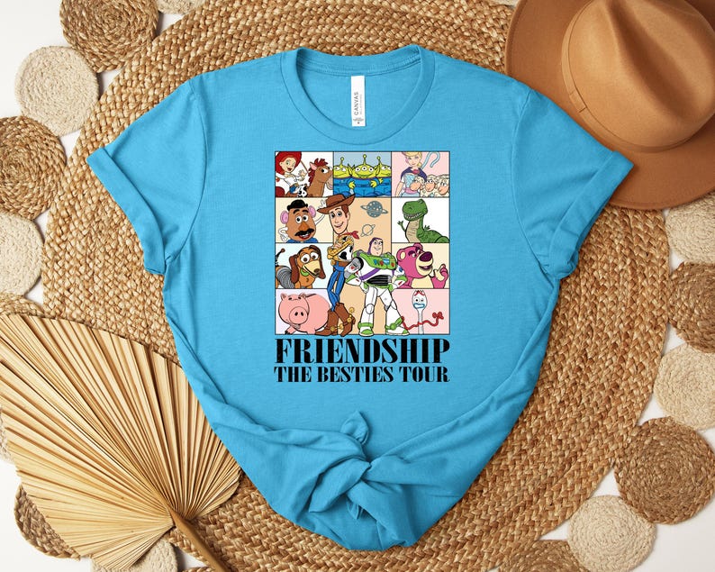 Puede incluir: Una camiseta turquesa con un gr&aacute;fico colorido de personajes de dibujos animados en una cuadr&iacute;cula. El gr&aacute;fico incluye personajes como Woody y Buzz Lightyear. El texto "FRIENDSHIP THE BESTIES TOUR" est&aacute; impreso debajo.