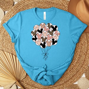 May include: A light blue t-shirt featuring a graphic of Mickey Mouse head-shaped balloons in black, pink, and leopard print. The balloons are tied together with a black ribbon. The shirt is made of a soft material.
