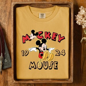 May include: Mustard yellow sweatshirt with a Mickey Mouse graphic. The design features the words "Mickey Mouse" in red and black, and the numbers "1924". The sweatshirt is presented in a wooden tray.