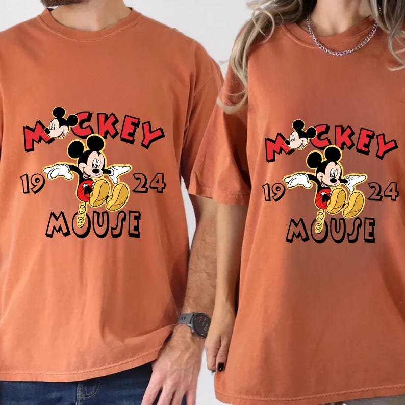 May include: Two rust-coloured t-shirts featuring a Mickey Mouse graphic. The graphic includes the words "Mickey Mouse 1924" in a playful font, with Mickey Mouse in the centre. The shirts are short-sleeved.