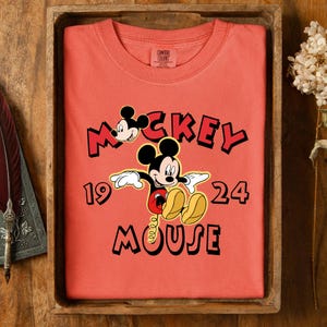 May include: Coral t-shirt with a Mickey Mouse graphic and the text "Mickey Mouse 1924". The design shows Mickey's face and full body in a playful pose. The shirt is folded neatly in a wooden tray, presented on a wooden surface.