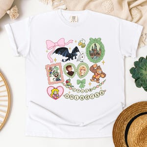 May include: White t-shirt featuring colorful cartoon illustrations of characters from various franchises. The design includes a dragon, castle, and characters from Mario, and other popular media. The text "EPIC UNIVERSE" is also included.