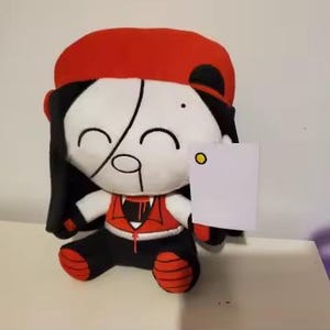 May include: A plush doll with a white face, black hair, and a red hat. The doll is wearing a red vest and black pants. The doll is holding a blank white card. The doll has a smiling expression.