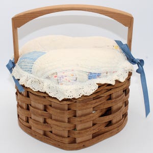 May include: A heart-shaped woven wood basket with a single arched handle. The basket features a quilted fabric liner with a white lace trim and blue ribbon ties on each side. The liner displays a light blue and white patchwork pattern.