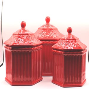 May include: A set of three bright red ceramic canisters with matching lids. Each canister features a vertical ribbed body and a hexagonal lid with a decorative embossed pattern and a small round knob handle on top.