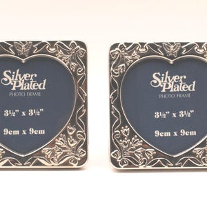 May include: Two square silver-plated photo frames with heart-shaped openings. The frames feature embossed butterfly, ribbon, and floral motifs. Text on the dark blue inserts reads: Silver Plated PHOTO FRAME 3 1/2" x 3 1/2" 9cm x 9cm.